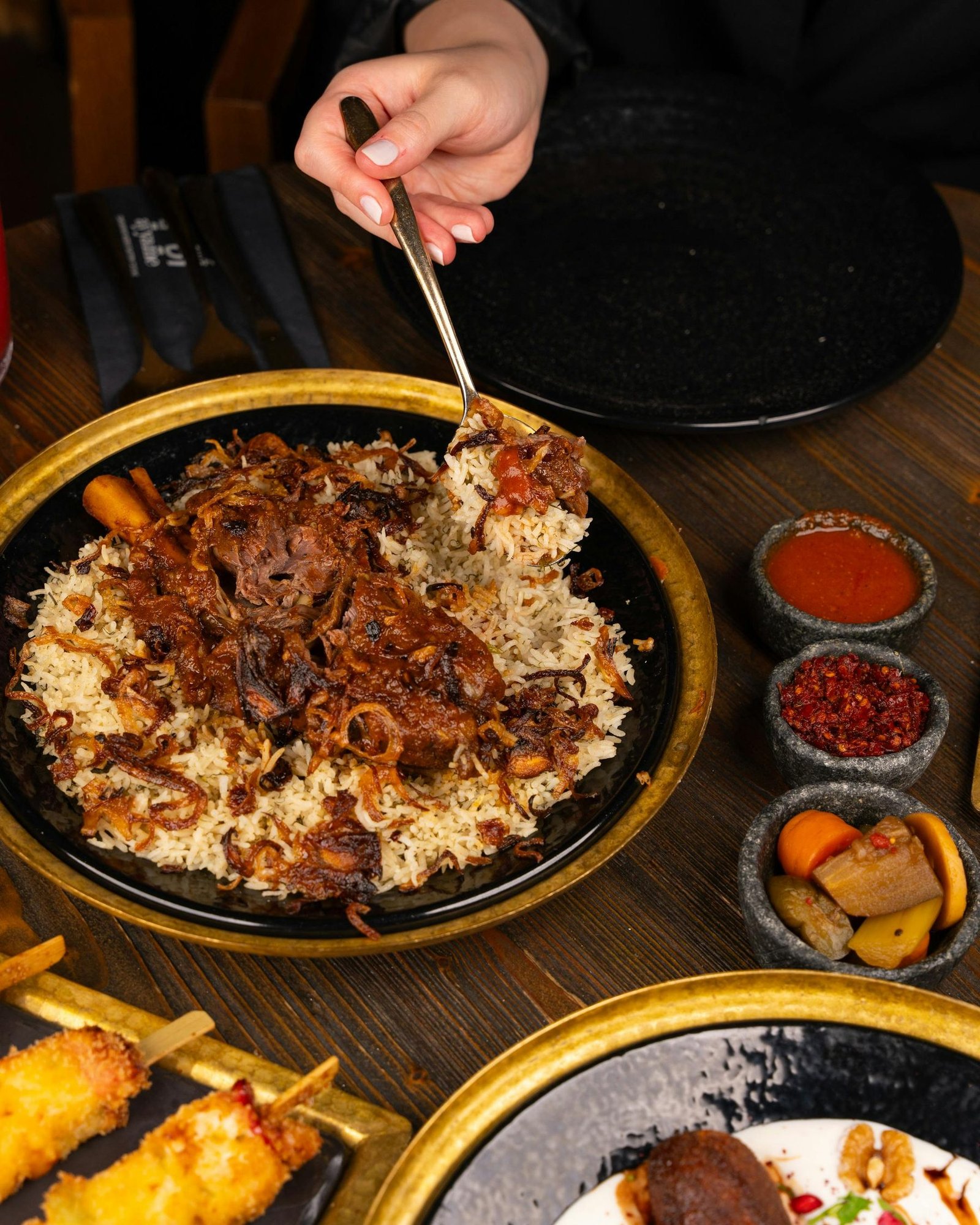 Kabsa-style lamb on a black plate with crispy onions, served with traditional Arabic sauces on the side